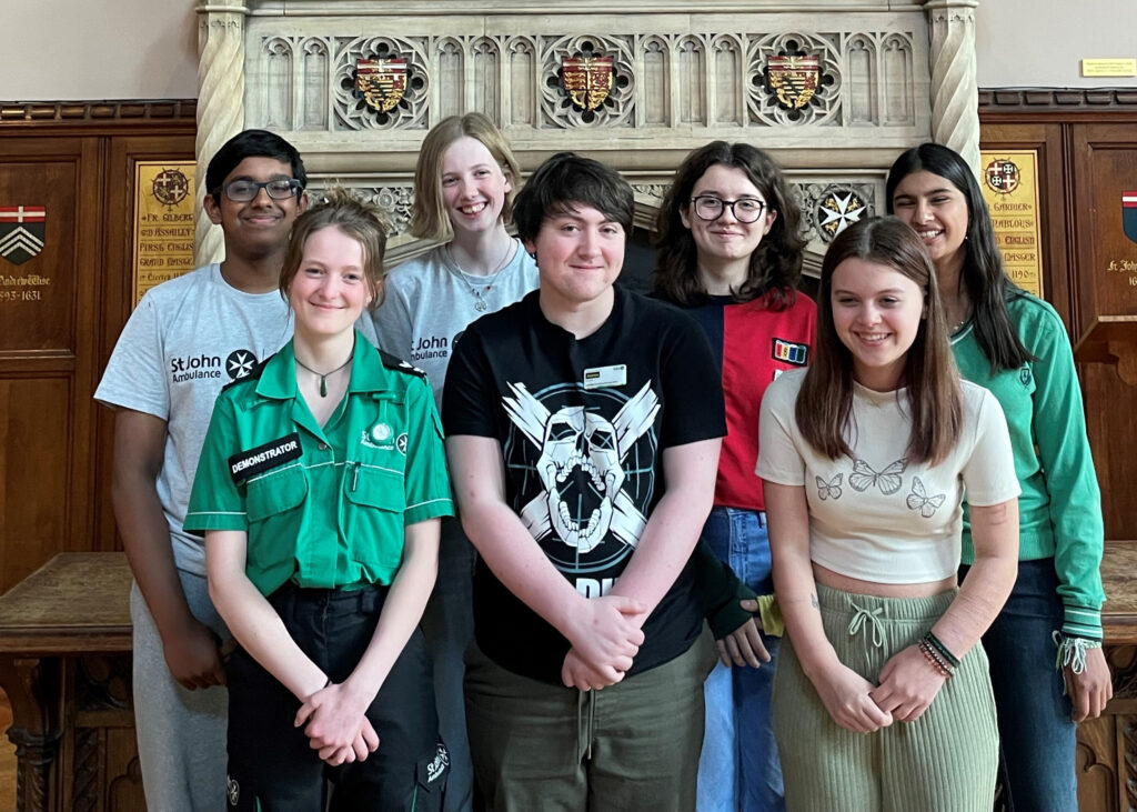 Colour photograph showing 7 Cadets standing in two rows in front of an elaborate stone fireplace. They are all smiling at the camera.