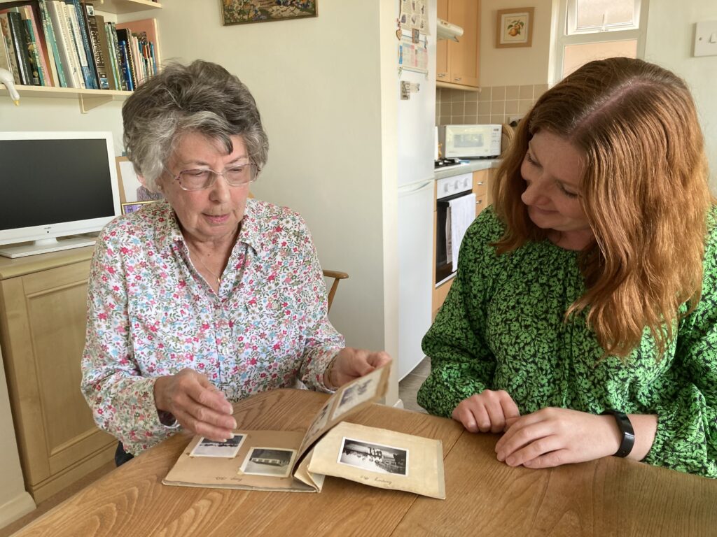 Colour photograph of an older woman sitting at a table with an open photograph album in front of her. She is showing it to a younger woman who is looking at the photographs and smiling. They are sitting in a dining room with kitchen visible beyond.