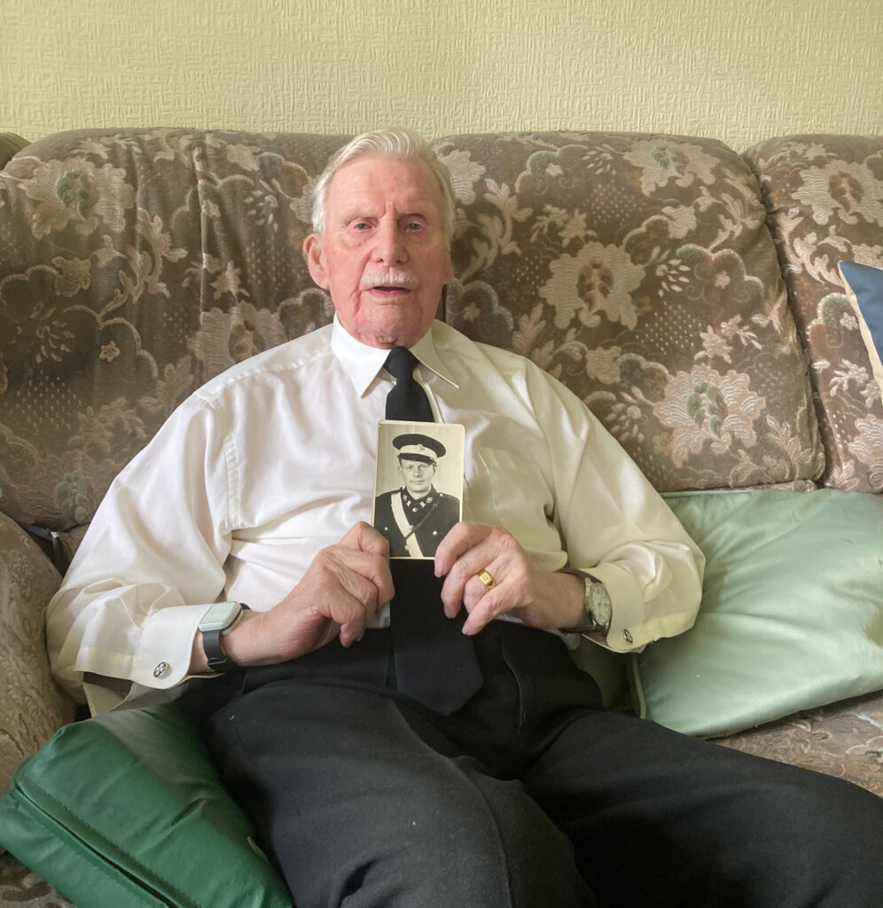Colour photograph of an elderly man sitting on a brown sofa and holding a black and white photograph of himself as a sixteen year-old out in front of him with both hands. He is wearing a white shirt, black tie and black trousers.