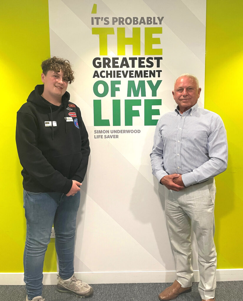 Colour photograph of a Cadet standing on the left wearing jeans and a black hoodie. Next to him stands an older man wearing cream trousers and a light blue shirt. They are posing against a wall graphic which has a quote reading &apos;It&apos;s probably the greatest achievement of my life&apos; Simon Underwood Life Saver.