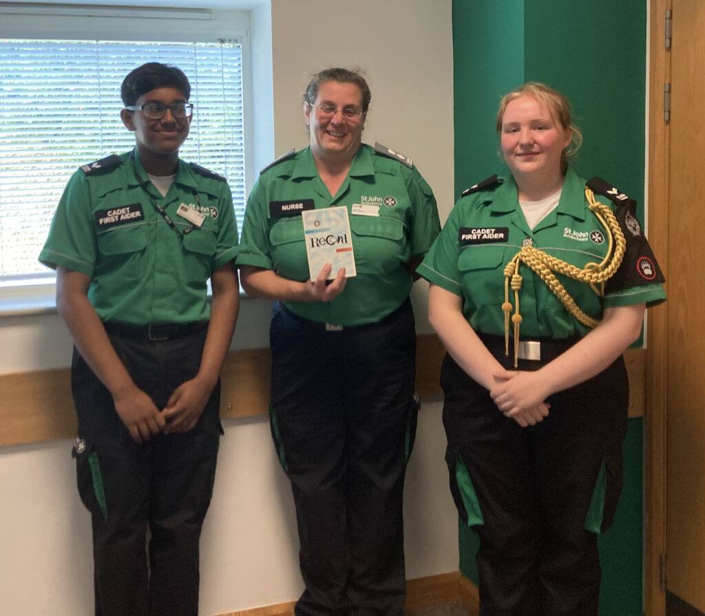 Colour photograph of two Cadets in green and black St John uniform standing either side of a woman, also in green and black St John uniform. They are inside a training room and the woman holds up a small booklet which has a title reading &apos;The Cadet Record Book&apos;.