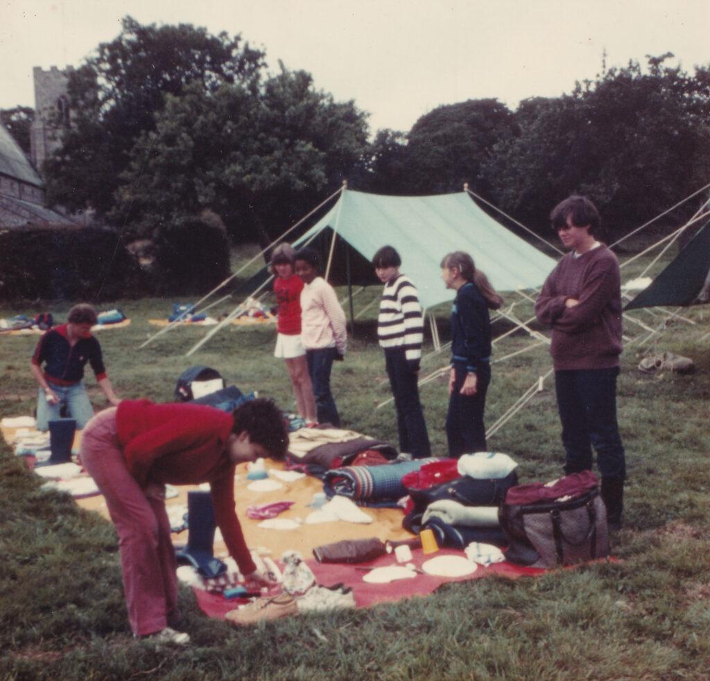 Colour photograph of seven young people standing around items of camping equipment that are laid out on a blanket in a field. Behind them are two tents.