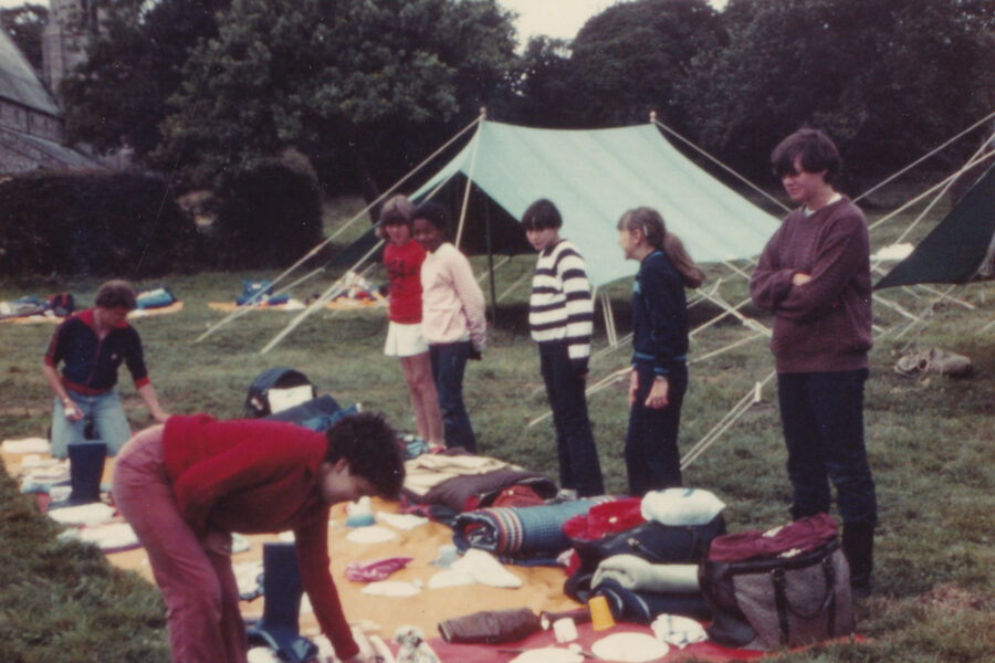 Colour photograph of seven young people standing around items of camping equipment that are laid out on a blanket in a field. Behind them are two tents.