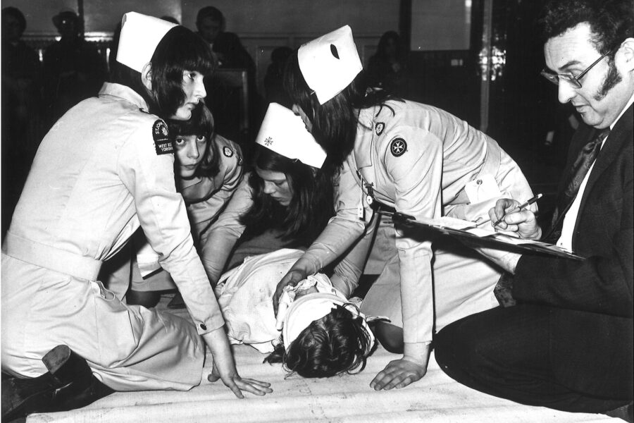 Black and white photograph of four female Cadets in uniform treating a fifth girl who is lying on the floor as a casulty simulation. An adult in uniform is looking on and making notes on a clipboard.