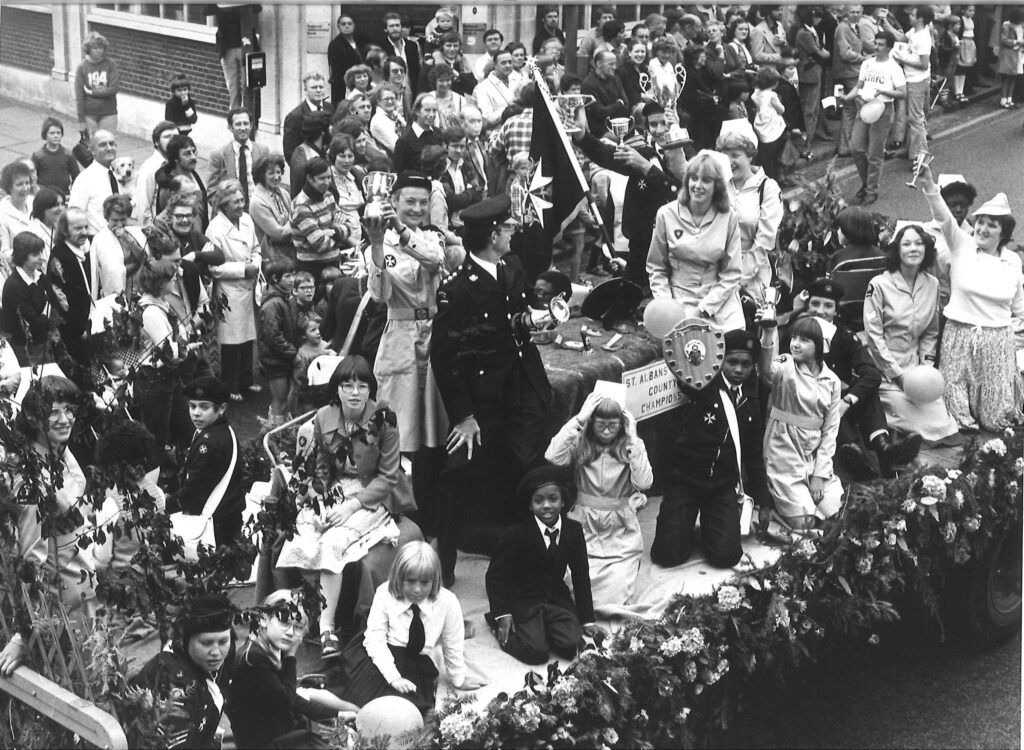 Black and white photograph of a group of young Cadets and adults in uniform on a carnival parade float. The Cadets hold up trophies. Behind them is a crowd of onlooking lining the road.