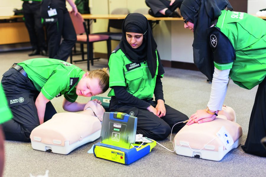 Colour photograph of three female Cadets in St John Ambulance uniform kneeling on the floor of a hall. Two of the Cadets are conducting First Aid on a dummy while the middle Cadets looks on.