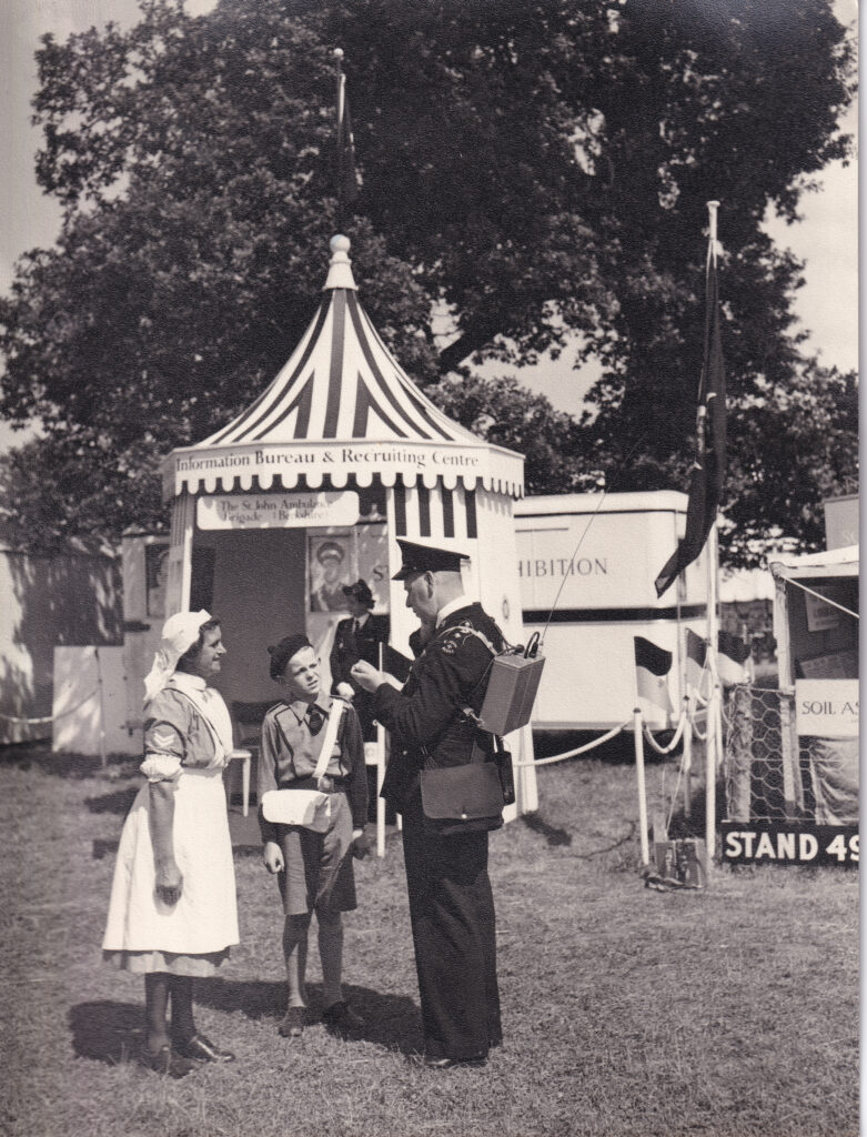 Black and white image of three people standing in front of a striped tent. On the tent is a sign that reads Information Bureau and Recruiting Centre. The person on the left is a nurse in uniform, wearing a white pinafore over a dress and a white headscarft. The person is the centre is a young boy in a Cadet uniform of shorts and shirt. The person on the right is a man in a black suit and hat. He is carrying a communication device on his back.