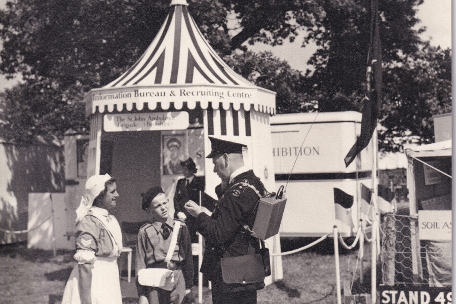 Black and white image of three people standing in front of a striped tent. On the tent is a sign that reads Information Bureau and Recruiting Centre. The person on the left is a nurse in uniform, wearing a white pinafore over a dress and a white headscarft. The person is the centre is a young boy in a Cadet uniform of shorts and shirt. The person on the right is a man in a black suit and hat. He is carrying a communication device on his back.