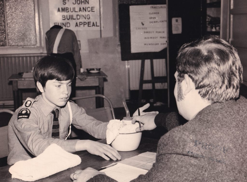 Black and white photograph of a young boy in uniform sitting at a table across from an adult male in a suit. The photograph is taken from behind the adult and so his face isn't shown. In front of the boy is a bowl of medical supplies. In the background there is a sign on the wall which reads St John Building Fund Appeal