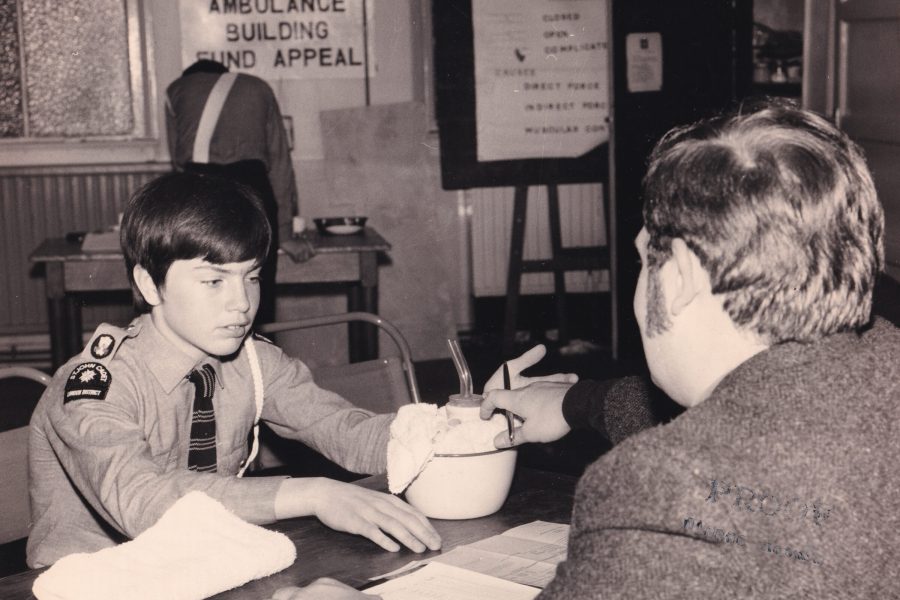 Black and white photograph of a young boy in uniform sitting at a table across from an adult male in a suit. The photograph is taken from behind the adult and so his face isn't shown. In front of the boy is a bowl of medical supplies. In the background there is a sign on the wall which reads St John Building Fund Appeal