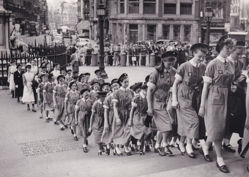 Black and white photograph of a large group of female nurses in uniform walking up the steps of St Paul's Cathedral. Behind them, a small crowd of onlookers stand at the roadside.