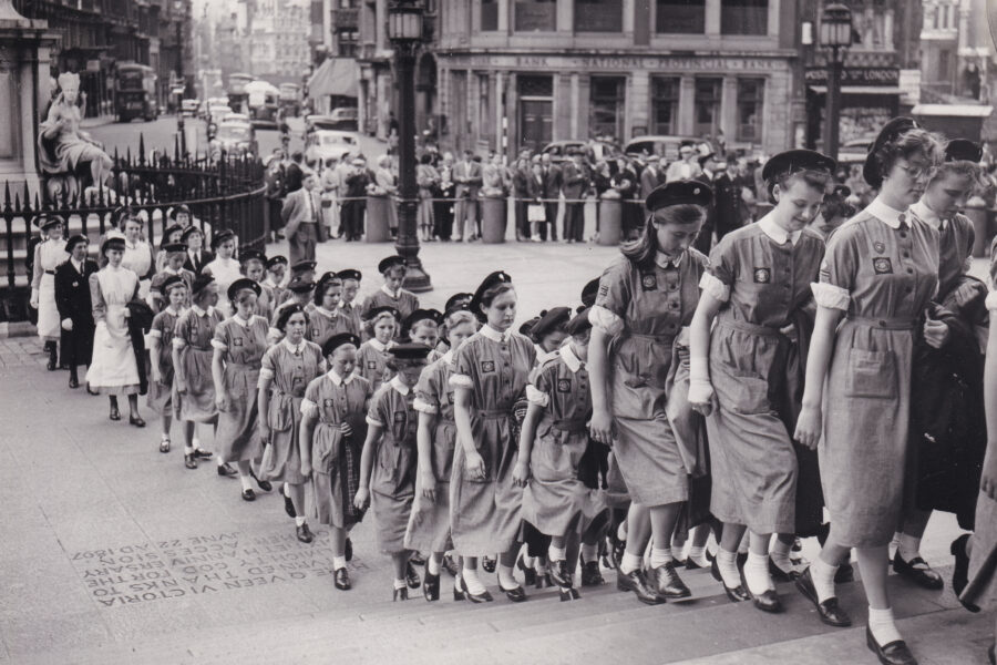 Black and white photograph of a large group of female nurses in uniform walking up the steps of St Paul's Cathedral. Behind them, a small crowd of onlookers stand at the roadside.