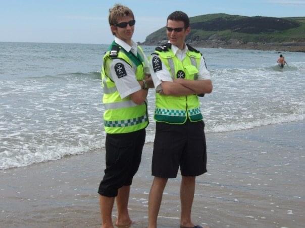 Colour photograph of two teenage boys standing on a beach. They are wearing sunglasses and St John Ambulance high vis jackets with black shorts.