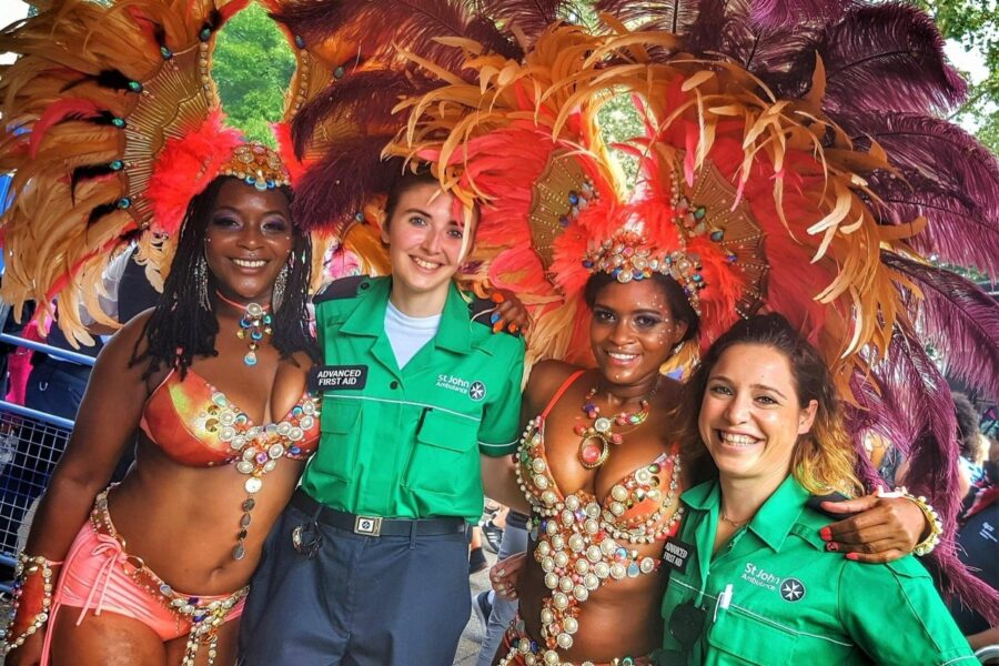 Colour photograph of two women in carnival costumes standing with their arms around two female St John Ambulance Cadets in uniform.