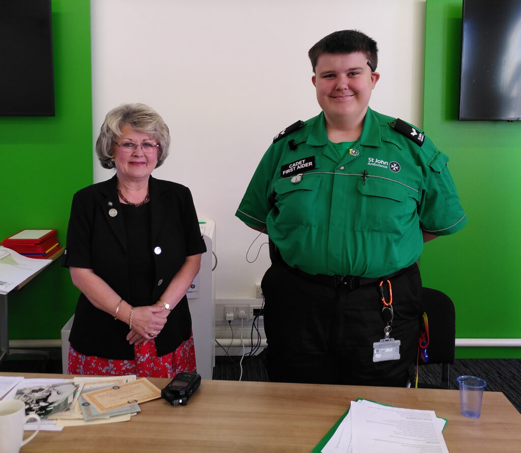 Colour photograph of a woman wearing a black jacket and red skirt standing next to a male Cadet in green and black St John uniform. A black recorder is on the table in front of them but the interview is not in progress. They are posing for a photograph and smiling at the camera.