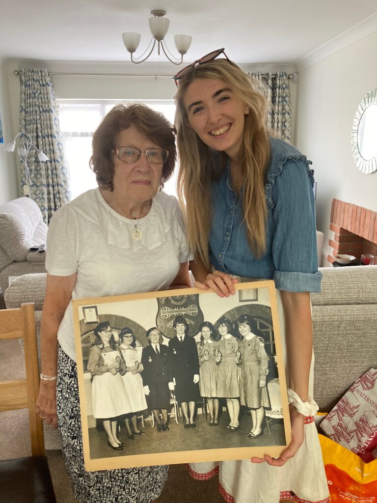 Colour photograph of an older woman wearing a white top and patterned skirt. She is standing next to a younger women and they are posing for a photograph in a living room. The younger woman holds a large black and white photograph showing a group of women and girls in St John uniform.