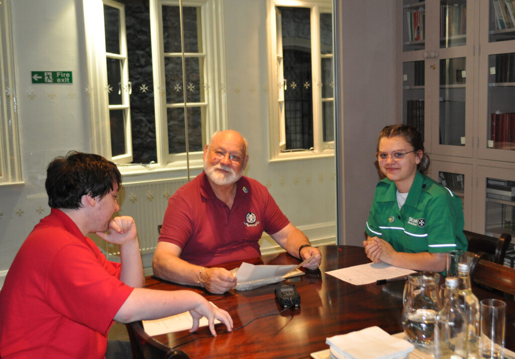 Colour photograph of an older man and two Cadets seated around a polished wooden table. There is a black audio recorder on the table. They all have paperwork in front of them. The older man and one of the Cadets are smiling at the camera.