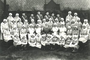 A black and white formal photograph of the Rochford girl and boy Cadet Divisions taken outside. In the foreground a row of girls dressed in white nursing caps and grey dresses sit on the floor, behind them is another row of girl Cadets and female adult members seated, behind them are two rows of standing Cadets. Of the 45 people photographed, only six are boys. They stand in the back row wearing black berets and grey shirts.