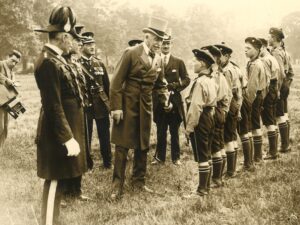 Sepia photograph showing 7 male Cadets on the right standing in a park and being inspected by the Duke of Connaught who wears a top hat and holds a walking stick. The Duke is accompanied by a group of five other men and a male photographer is just visible to the far left holding his camera.