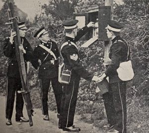 Four men in St John Ambulance Brigade uniform are standing by the side of the road. One of the men is holding a stretcher. Two of the men are replenishing supplies of a first aid box which is attached to a pole at the side of the road. The fourth man is watching on.