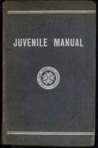 A colour image of the front cover of the 1929 Juvenile Manual. It is a blue material cover with white imprinted wording 'JUVENILE MANUAL' and the white St John Ambulance Association circular logo featuring the eight-pointed cross.