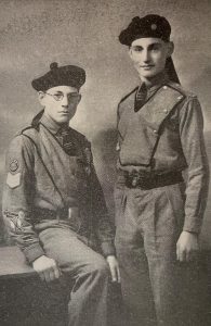 A black and white photograph of two boys in Cadet uniform. The boy on the left it perched on a table, and the boy on the right is standing up.