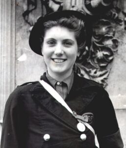 Black and white photograph of a young woman smiling directly at the camera. She wears a black hat, grey shirt and dark jacket with an ARP badge stiched to her left pocket. The George Medal is pinned to her jacket.
