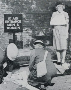 Black and white image of a Nursing Cadet wearing an arm sling, in front of her two personnel attend to simulated casualties on stretchers