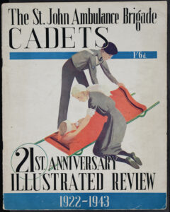 Colour photograph of the magazine cover which reads &apos;The St John Ambulance Brigade Cadets. 21st Anniversary Illustrated Review. 1922-1943. 1/6d&apos;. The illustration shows a Nursing Cadet and an Ambulance Cadet in their grey St John uniforms tending to a patient lying on a stretcher with a bandage around his head and covered in a red blanket. The illustration appears against a white background.