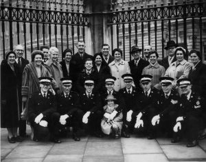 A group of men and women posing for a photograph outside the gates of Buckingham Palace. Kneeling on the front row are uniformed male members of St John Ambulance, and standing behind are friends and family.