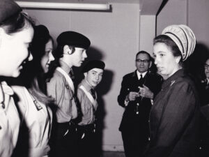 Black and white photograph of a young Princess Anne standing on the right and wearing a white hat and a dark jacket. The Princess looks towards a group of Cadets, two girls and two boys, wearing St John uniform.