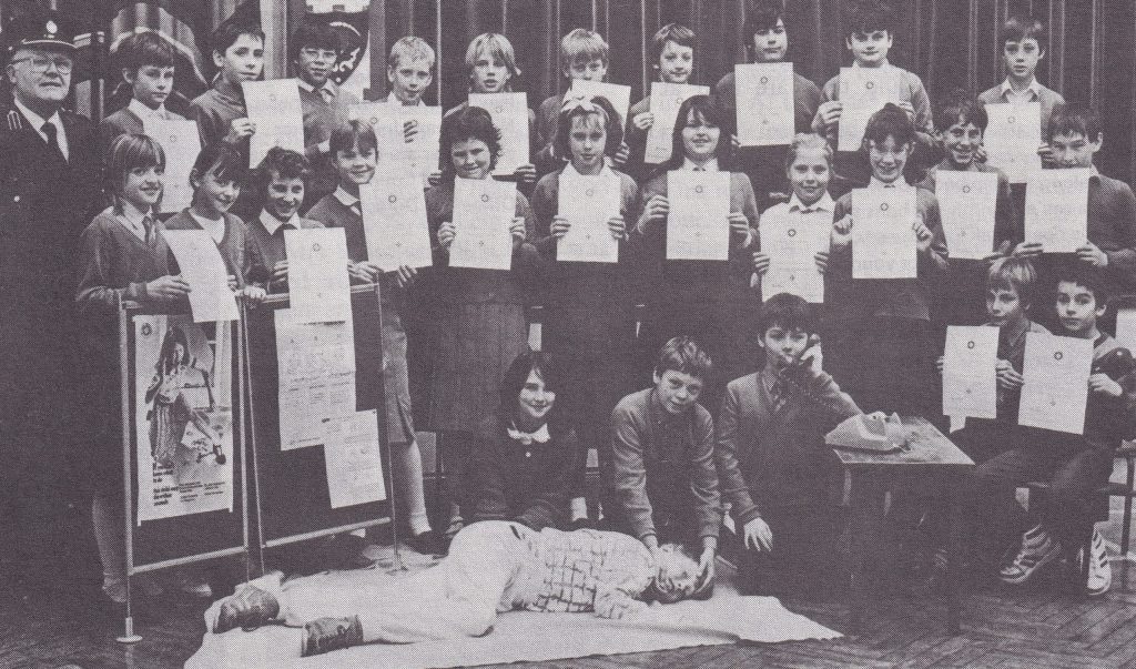 A group of children line up holding certificates. Some children are demonstrating putting a casualty into the recovery position and calling for help.