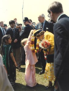 Queen Elizabeth II dressed in yellow, bows slightly to receive a floral garland from a young child, while adults watch.