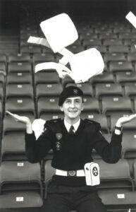 Seated male Cadet smiles as he throws two white haversack bags into the air above his head. He wears a black beret, black jumper, black trousers, white belt and white first aid pouch attached to the belt and decorated with a black 8-pointed cross.