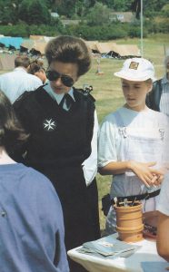 Colour photograph of Princess Anne wearing black and white St John uniform and sunglasses stands in front of a table where a Cadet activity is taking place. Tents are visible in the field beyond. Next to the Princess is a female Cadet wearing dungarees.