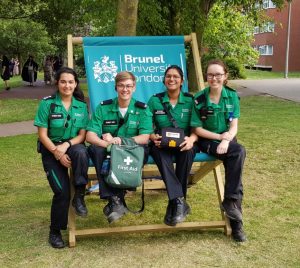 Colour photograph of four St John Ambulance student members in green and black uniform, seated on an oversize deckchair printed with Brunel University logo