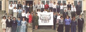 Colour photograph of 41 members of the first National Youth Council standing on the stone steps in front of a manor house. Princess Anne stands at the centre next to a huge black and white illustration of the National Youth Council logo, being held up by two members. The logo features two hands clasped together above a banner than reads National Youth Council and an eight-pointed cross.
