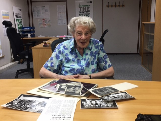 Colour photograph of an older woman sitting behind a table. She is looking directly at the camera. In front of her there is a pile of black and white photographs on the table.