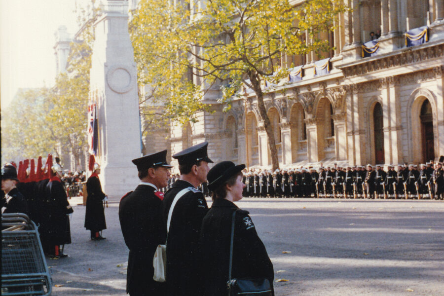 Colour photograph of three people in St John uniform lining the roadside. Behind them is the cenotaph along with lines of other people in official uniforms.