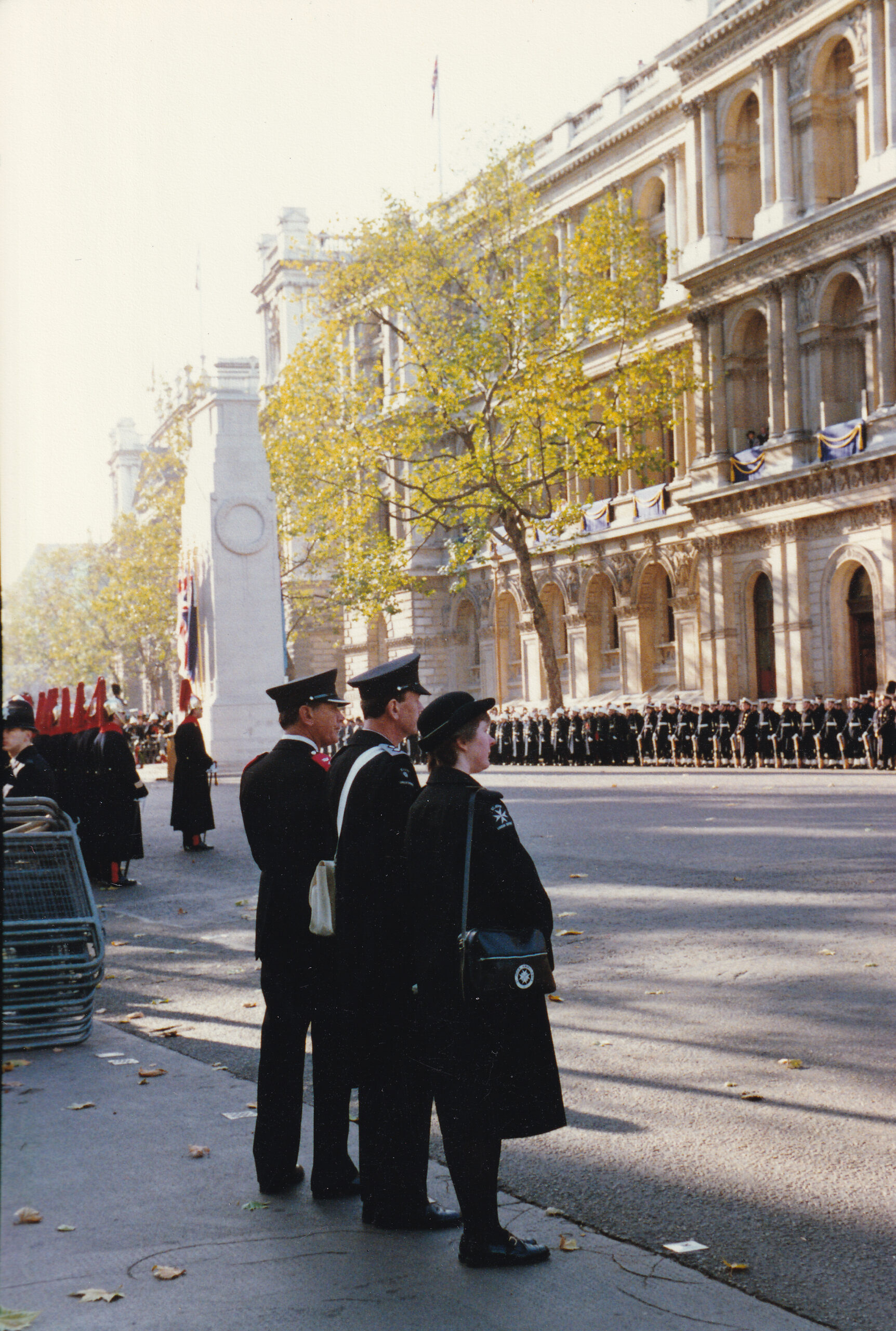 Colour photograph of three people in St John uniform lining the roadside. Behind them is the cenotaph along with lines of other people in official uniforms.