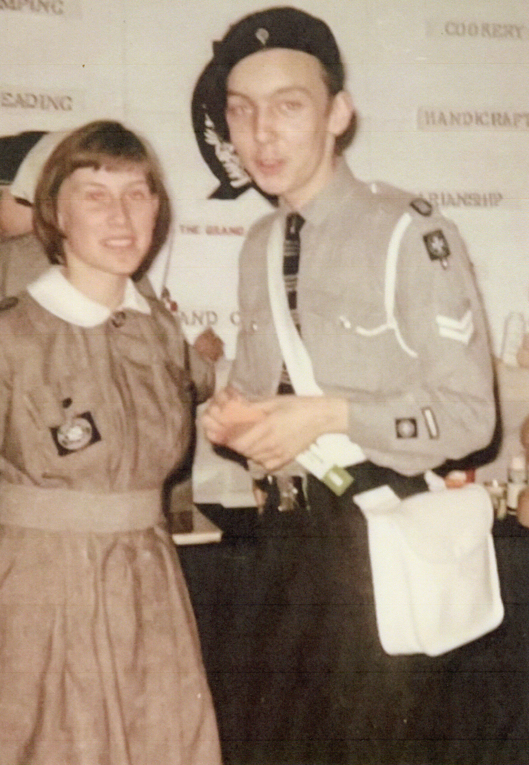A faded photograph with a white Nursing Cadet on the left wearing a grey knee length dress with a white collar belted at the waist. The dress has a pocket on the chest with a black badge bearing St John’s 8 pointed cross. She is also wearing a white nursing cap towards the back of her head. To the right of the photo is a white male Cadet wearing a black beret