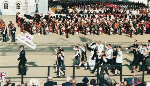 Colour photograph of St John Ambulance members taking part in a parade and marching past Horse Guards where a band is playing and onlookers are seated in stands to watch. The march is led by a male member of St John in formal black uniform holding a placard that reads '100 St John Ambulance'. He is followed by St John Ambulance youngest members, known as Badgers, wearing their black and white uniforms and accompanied by an adult in the Bertie Badger costume and they are all waving to the crowds. Adult and Cadet members march behind.