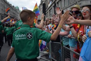 A young Cadet in green and black St John uniform takes part in the parade. They have their back to the camera and are high-fiving people in the crowd. In front of them are other members of St John Ambulance waving rainbow flags.