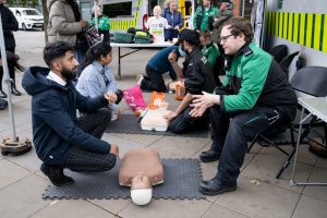 In the foreground is a seated and uniformed member of St John Ambulance, talking to a member of the public opposite him. In-between them on the ground is a resuscitation Annie mannequin. In the background, other uniformed members of St John Ambulance are showing other members of the public how to perform CPR.