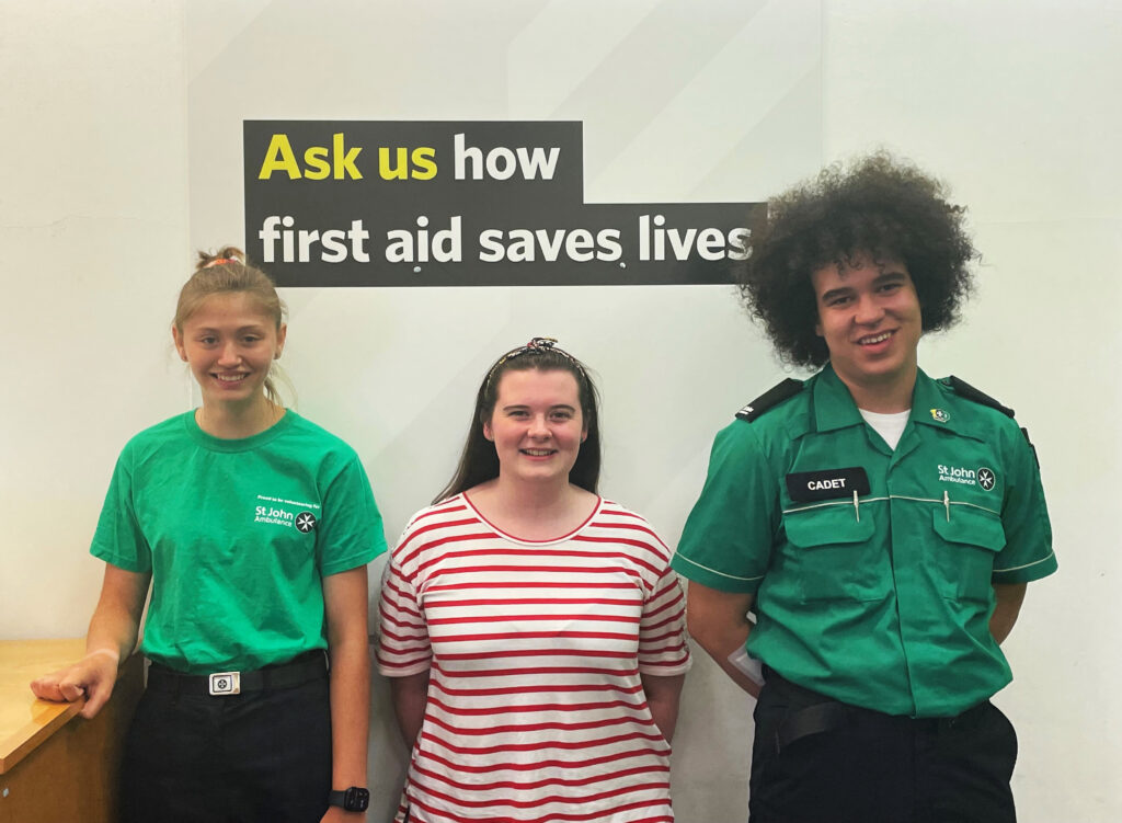 Colour photograph of two Cadets and a young woman standing in a room beneath a wall graphic that says &apos;Ask Us How First Aid Saves Lives&apos;. The Cadets are both wearing green and black uniform, the woman is wearing a red and white striped top. They are all smiling at the camera.