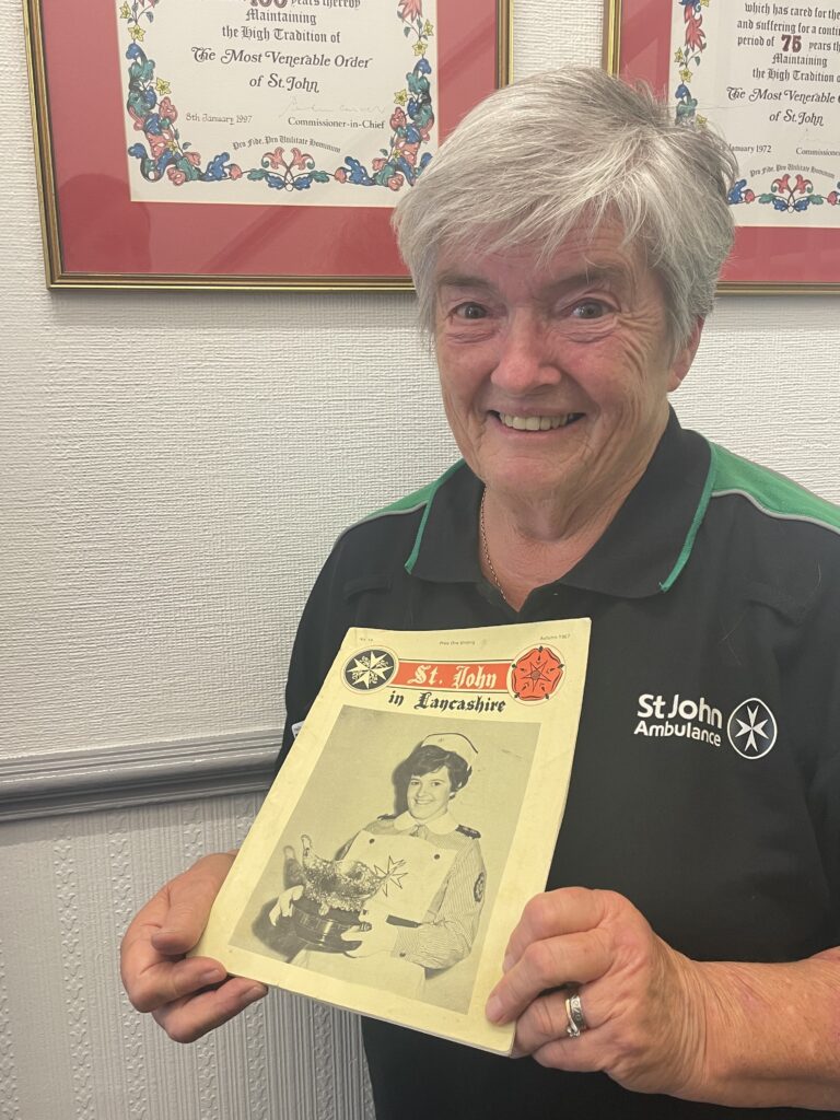 Colour photograph of an older lady wearing a green and black St John t-shirt. She is smiling and holding up an old magazine that has a black and white image of her on the cover as a young woman.