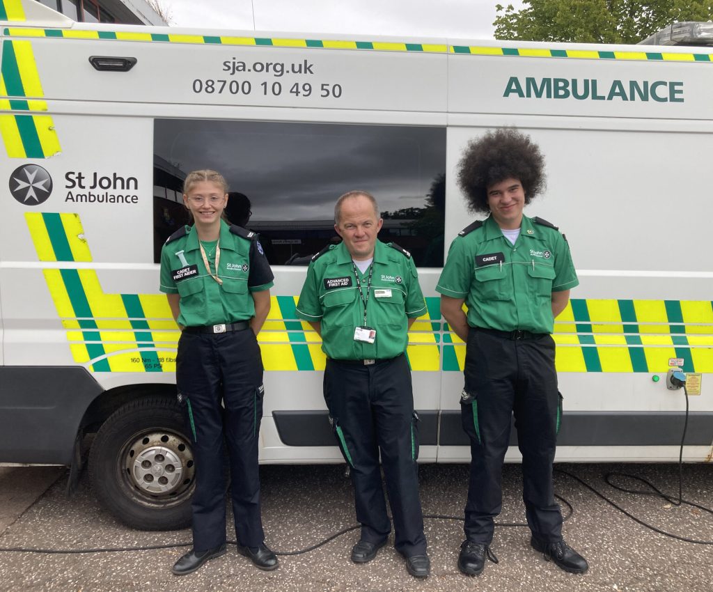Colour photograph of a man and two Cadets standing outside in front of a white St John ambulance with luminous yellow and green chevrons. All three are wearing green and black St John uniform and smiling at the camera.