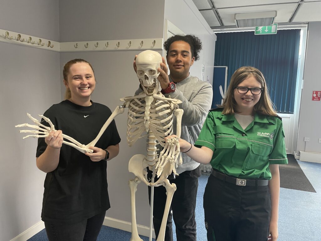 Colour photograph of a young woman and two Cadets standing in a room. They are posing for a photograph around a plastic teaching skeleton. They are all looking at the camera and smiling.