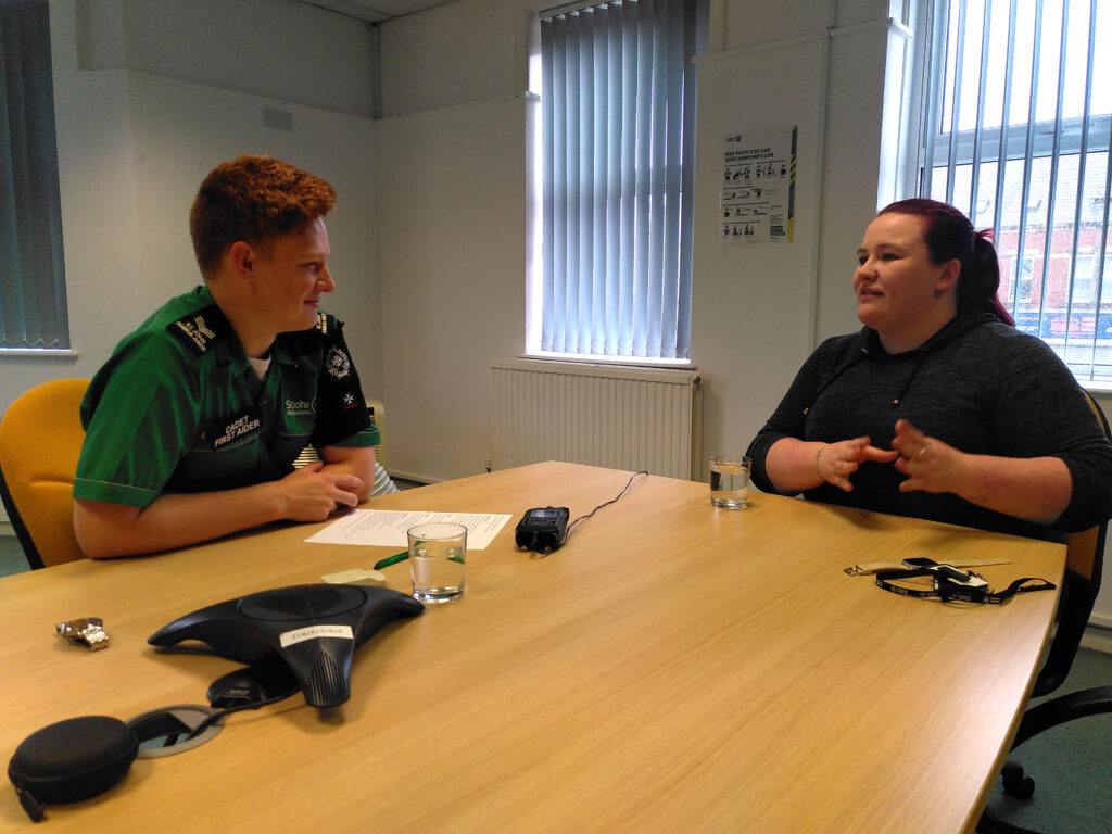 Colour photograph of a male Cadet and a woman seated at a table. An interview is in progress and there is a black audio recorder on the table between them. The woman is gesticulating with her hands as she explains something.