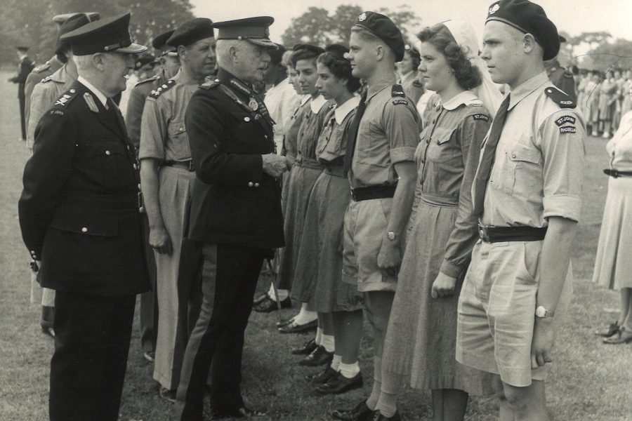 Black and white photograph of male and female Cadets in uniform standing to attention in a field. They are being spoken to by a group of adult males in uniform.